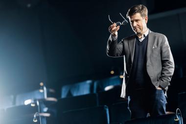 Kuvassa on oikealla Klaus Härö seisomassa ja puhumassa lavalla, jossa on taustalla istuinrivejä. Hänellä on harmaa takki ja tumman sininen liivi. Hän pitää silmälaseja oikeassa kädessään ylhäällä. Tausta on tumman sininen ja vasemmalle heijastuu valoa.