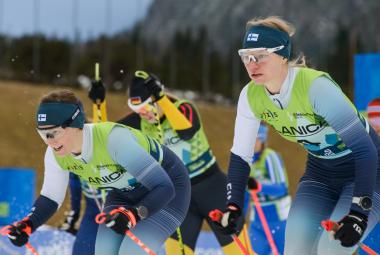 Kuvassa on kolme naishiihtäjää lähdössä kilpailemaan. Heillä on Suomen sini-valkoiset edustusasut ja pipot, joissa on Suomen lippu. 