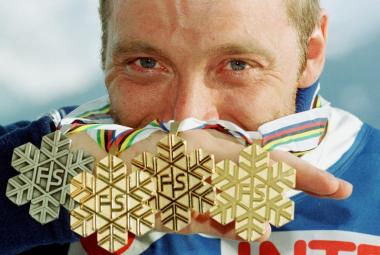 Kuvassa on taustalla Mika Myllylän kasvot ja hän pitää puoliksi kasvojen edessä neljä mitalia, joissa on havukuviota ja keskellä kirjaimet FS. taustalla vaalean sinistä pilveä.