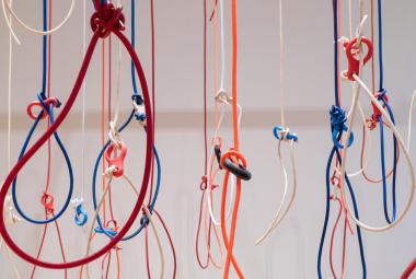 Kuvassa on roikkumassa erivärisiä naruja ja niissä on solmuja. Joihinkin on solmittu iso silmukka. Tausta on harmaa.