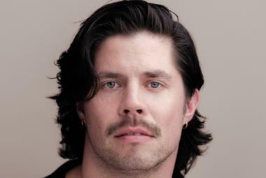 Kuvassa on Markku Haussilan kasvokuva ja hänellä on tummat puolipitkät hiukset. Tausta on harmaa.