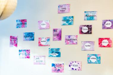 Kuvassa on vaalean sinisellä ja vaalean keltaisella pohjalla lappuja seinällä ja niiden värit ovat puna-sini-valkoisia.  Niissä on tekstejä kuten kiva, rohkea, herkkä jne.