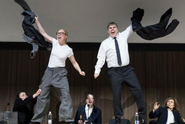 Kuvassa on nuori nainen vasemmalla ja hän heiluttaa takkiaan ja hänellä on valkoinen t-paita ja harmaat, löysät housut päällää, Oikealla on nuori mies, jolla on valkoinen paita jamusta kravatti sekä mustat housut päällään. Taustalla alhaalla on kolme henkilöä pöydän ääressä mustissa takeissa.