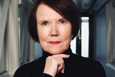 Kuvassa on Rosa Liksom puolivartalokuvassa mustassa poolopuserossa ja hänellä on oikea käsi leuan alla. Hänellä on tummat, punertavat puolipitkät hiukset. Taustalla on harmaata käytävää.