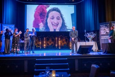 Kuvassa on sinisävyinen näyttämö ja vasemmalla seisoo viisi henkilöä taputtamassa. Oikealla seisoo mies, joka puhuu. Taustalla näkyy ylhäällä valkokangas ja siinä on naisen kasvot. Hän pitää käsiään poskillaan.