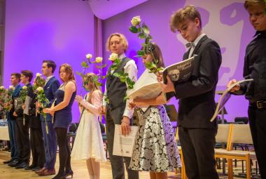 Kuvassa on viistosti seisomassa näyttämöllä sekä tyttöjä että poikia. Heillä on kädessään ruusu ja kunniakirja. Taustalla on violettia valoa.