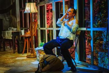 Kuvassa on Ilja Peltonen kalustetussa huoneessa, jossa Ilja Peltonen istuu tuolilla ja nojaa taaksepäin.  Hänellä on vaalean sininen paita ja siniset housut päällään. Huoneessa on pieni pöytä ja iso jalkalamppu sekä takana isot ikkunat. 