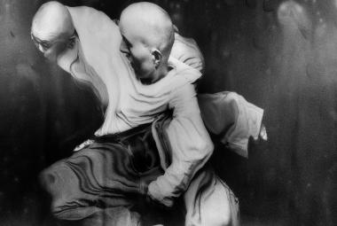 Kuvassa on kaksi miestanssijaa, jotka ovat kietoutuneitä toisiinsa vaalealla paidalla. Heillä on tummat housut. Kuva on musta-valkoinen.