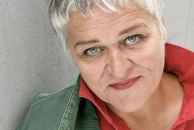 Kuvassa on Pirkko Saision kasvokuva ja hänellä näkyy kuvassa harmaata takkia ja punaisia kauluksia. Hänellä on vaalean harmaat hiukset.