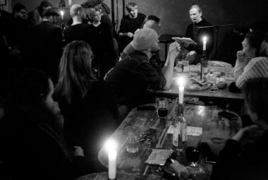 Kuvassa on huone täynnä ihmisiä. Oikealla on pitkä pöytä ja sen äääressä on ihmisiä juomassa ja keskustelemassa. Pöydässä on kynttilöitä. Vasemmalla on ihmisiä seisomassa. Kuva on musta-valkoinen.