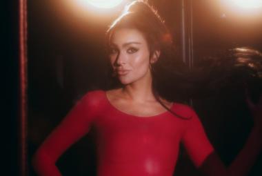 Kuvassa on keskellä Sara Siipola puolivartalokuvassa. Hänellä on punainen mekko päällään ja hänellä on tummat hiukset nutturalla. Taustalla tumman ruskeaa ja kaksi ylävaloa.