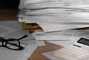 Kuvassa on iso pino valkoisia papereita ja vasemmalla on silmälasit ja oikealla edessä on valkoinen laskin. 