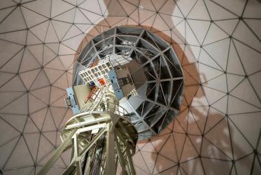 Kuvassa on avaruusteleskooppi kuvattuna alhaalta ylöspäin ja taustallaon rautaverkkoa vaaleanruskealla pohjalla.