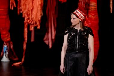 Kuvassa on roikkuvia tumman punaisia tilataideteoksia tummalla taustalla. Oikealla edessä on Elle Sofe Sara seisomassa ja hänellä on musta hihaton mekko ja punainen kirjailtu Lapin päähine päässään. 