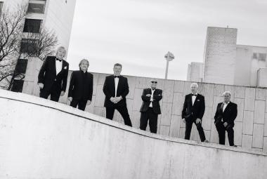 Kuvassa on alaspäin viettävän kaiteen vierellä kuusi mieslaulajaa seisomassa mustissa puvuissa.  Taustalla on vaaleaa seinämää ja vasemmalla kerrostalon julkisivua ikkunoineen. Kuva on musta-valkoinen.