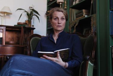 Kuvassa on Jonna Järnefelt istumassa nojatuolissa ja hänellä on kädessään muistikirja kädessään. Takana on ruskea matala kirjahylly ja oikealla on iso tumman ruskea kirjahylly.