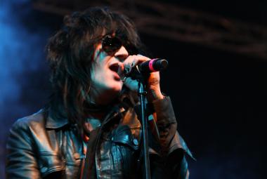 Kuvassa on Jyrki60 laulamassa nahkatakissa ja mikki kädessään. Kuva on puolivartalokuva ja tausta on tumma, vasemmalta hieman sininen. 