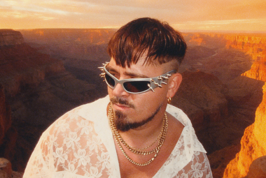 Kuvassa on Käärijä keskellä alempana ja hänellä on valkoinen paita päällään ja aurinkolasit. Taustalla on ilta-auringon valossa olevaa Grand Canonia.