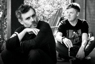 Kuvassa on vasemmalla edessä istumassa kyyryssä Gabriel Kahane ja hänen takanaan oikealla kyykyssä on Pekka Kuusisto. Taustalla seinää ja pensaistoa. Kuva on musta-valkoinen.