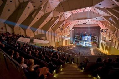 Kuvassa on Tampere-talon konserttisali ylhäältä alaspäin ja hämärässä salissa on yleisöä istumassa  ja katsomassa alhaalla olevalle näyttämölle orkesteria. Vasemmalla ylhäällä on taitettua kattoa.