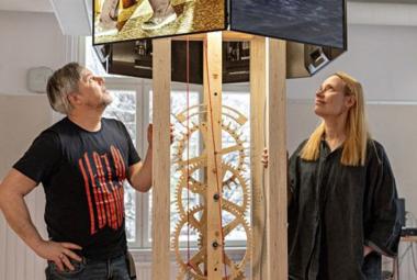 Kuvassa on vasemmalla Oliver Kochta-Kalleinen katsomassa ylöspäin puisen kellokoneiston yläpuolella olevaa neliötä ja sen koneistoa. Oikealla on Tellervo Kalleinen seisomassa ja katsomassa myös ylöspäin. Hänellä on musta leveä mekko. Taustalla on vaalealla seinällä ikkunoita.