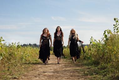 Kuvassa ovat Rodeon kolme jäsentä kävelyllä hiekkatiellä ja vierellä on korkeita kasveja pellolla. Heillä tummat pitkät mekot päällään. Taustalla on sinistä taivasta.