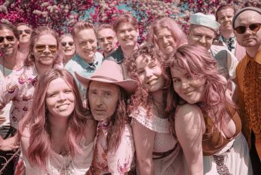 Kuvassa on puolivartalokuvassa runsaasti nuoria esiintyjiä tiiviisti vierekkäin ryhmänä koko kuva-alalla. Kuvan sävy on vaalean punainen.