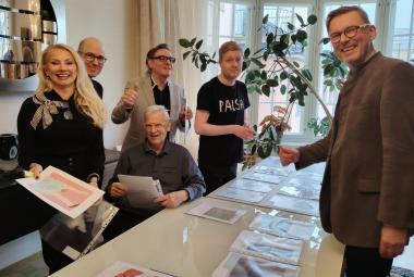 Kuvassa ovat suuren pöydän ääressä seisomassa on vasemmalla viisi henkilöä ja edessä oikealla seisoo yksi mies. Pöydällä on taideteosten luonnoksia A4 koossa.