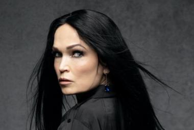 Kuvassa on Tarja Turunen puolivartalokuvassa ja hän katsoo olkapään yli. Hänellä on pitkät, tummat hiukset ja musta jakku. Tausta on tumman harmaa.