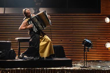 Kuvassa on vasemmalla jalustalla sivuttain Kimmo Pohjonen keltaisissa leveissä housuissa soittamassa harmonikkaa. Takana on ruskeaa vaakasuoraa rimaa.