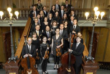 Kuvassa on Vaasan kaupunginorkesterin jäsenet seisomassa leveillä portailla ja taustalla sekä sivuilla on keskiruskeaa panelointi. Ylhäällä on molemmin puolin kolmen lampun valaisimet. 