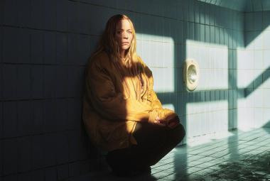   Kuvassa on Paula Vesala kyykussä ja vasen puoli kuvasta on tummaa ja oikealle tulee auringon valoa. Vesala näkyy puolittain. Taustalla on isojen lasi-ikkunoiden nurkka.