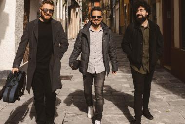 Kuvassa on kolme nuorta miestä kävelemässä kapeaa talojen reunustamaa katua. Heillä n mustat vaatteet paitsi keskimmäisellä harmaa paita.