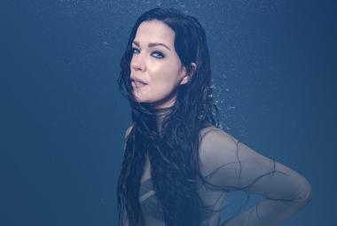 Kuvassa on sivuttain Jenni Vartiainen ja hänellä on tummat pitkät hiukset sekä hihoista läpinäkyvä pusero. Tausta on tumman sininen.