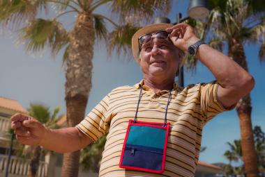 Kuvassa on Tom Lindholm kuvattuna yläviistoon. Hänellä on karttalaukku kaulassaan ja hän pitää aurinkolaseista kiinni vasemmalla kädellään. Taustalla on palmuja, vaaleita rakennuksia ja sinistä taivasta.