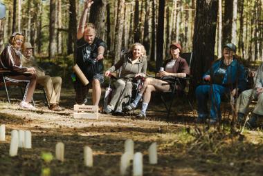 Kuvassa on metsässä istumassa nurmikon takana ja siinä näkyy miehiä ja naisia vasemmalla olevan karavaanerin nokka. Edessä on mölkkypalikoita pystyssä. 