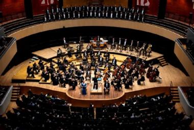 Kuvassa on Sinfonia Lahti ylhäältä kuvattuna soikealla puisella esiintymislavalla soittamassa. Ylempänä näkyy parvekkeella yleisöä ja edessä permannolla. Kuva on osa kirjan kannesta.