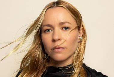  Kuvassa on Paula Vesala kasvokuvassa. Hänellä on pitkät, vaaleat hiukset ja ne liehuvat tuulessa. Tausta on vaalea.