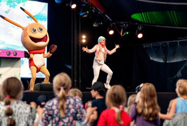 Kuvassa on esiintymislavalla vasemmalla Kikattava Kakkiainen, joka on oranssinen otus isoine päineen ja tuntosarvineen. Silla on punainen paita päällään. Lavalla oikealla on naislaulaja, jolla vaaleat vaatteet ja hän tekee loikkaa kädet levitettyinä.