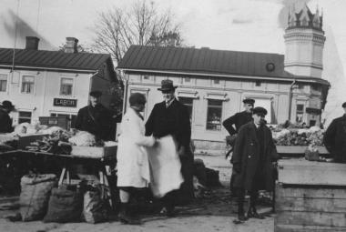 Kuvassa on etualalla torin myyntipöytä ja pöydän takana mies seisomassa. Pöydän vieressä seisoo nainen valkoisessa takissa ja mies mustassa hatussa ja lierihatussa. Oikealla on kaksi miestä ja välissä iso puulaatikko sekä sen vieressä mies. Taustalla on kaksi vaaleata, vanhaa puutaloa. Kuva on musta-valkoinen.