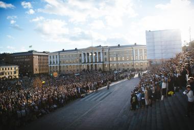 Kuvassa on Senaatintorilla vasemmalla runsaasti kuorolaisia seisomassa ja laulamassa. Oikealla on Helsingin tuomiokirkon portailla on myös runsaasti laulajia seisomassa. Keskellä on näkyvissä tilaa ja katukivetystä.