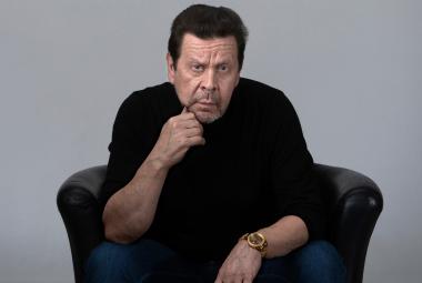Kuvassa on Pate Mustajärvi mustassa paidassa istumassa mustassa isossa nojatuolissa kumartuneena eteenpäin. Hän nojaa oikealla kädellään leukaansa. Tausta on harmaa.