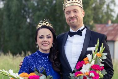 Kuvassa on vasemmalla Raija Mäntyniemi sinisessä mekossa ja vieressä oikealla Juho Punkeri mustassa takissa ja valkoisessa paidassa. Molemmilla on kukkakimput kädessään. Taustalla on metsää ja valkoinen talo.