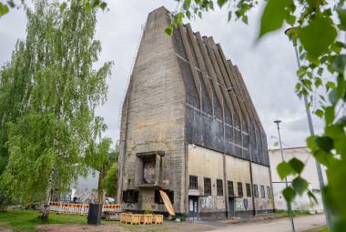 Kuvassa on keskellä harmaa, ylöspäin menevä harmaa siilo. Siinä on kattojuonteita tummalla katolla. Alhaalla on rivi ikkunoita. Molemmilla puolilla on vihreälehtisiä lehtipuita.