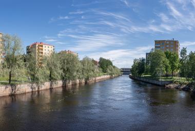 Kuvassa on keskellä joki ja sen molemmin puolin kerrostaloja, joiden edessä on lehtipuita. Takana on sinistä taivasta.