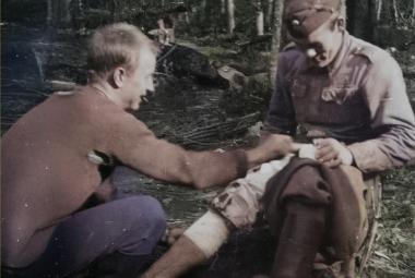 Kuvassa on metsikössä vasemmalla jalan sitomistilanne ja siinä vasemmalla on mies istumassa maassa ja pitää maassa olevan sotilaan sidotun jalan polvesta kiinni. Oikealla toisella puolella on sotilaspuvussa oleva mies tekemässä sidontaa.