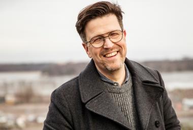 Kuvassa on Klaus Härö puolivartalokuvassa ja hänellä on ruskea takki ja sen alta näkyy harmaan ruskeata villapaitaa. Taustalla on epätarkkaa syysmaisemaa. 