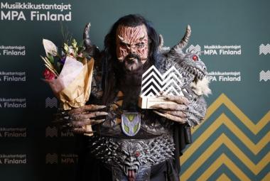 Kuvassa on Lordi monsteriasussaan, jossa on juovikas naamari ja olkapäistä lähtevät sarvet. Pukuun kuuluvat musta, paksu vyö, jossa on hirviön naama.  Käsissään Lordilla on kukat ja palkinto. 