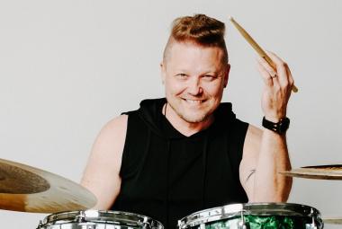 Kuvassa on Tomi Salesvuo hihattomassa mustassa hupparissa. Hän soittaa rumpuja. 