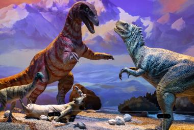 Kuvassa on kuvassa on vasemmalla violetti iso dinosaurus ja sitä vastapäätä on turkoosi iso dinosaurus. Punertavan dinosauruksen vieressä on pieni turkoosi dinosaurus. Maassa on kelopuu ja sen päässä on orava . Taustalla on sinertävän värikästä vuoristoa.